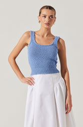 A woman is wearing a sleeveless, light blue, knitted crop top with wide straps and a ribbed hem. She pairs it with a high-waisted, white skirt that has a gathered, voluminous design. She is accessorized with gold hoop earrings and rings.